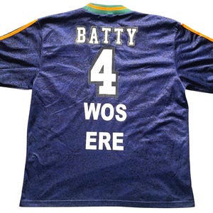 May include: A blue jersey with the name "BATTY" and the number "4" in white block letters. The jersey also has the words "WOS ERE" in white. The sleeves have green and orange accents.