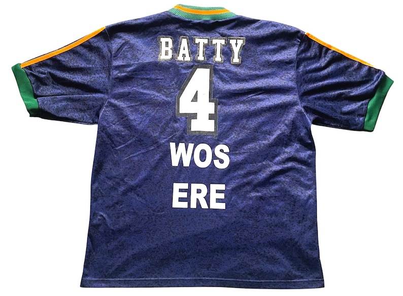 May include: A blue jersey with the name "BATTY" and the number "4" in white block letters. The jersey also has the words "WOS ERE" in white. The sleeves have green and orange accents.