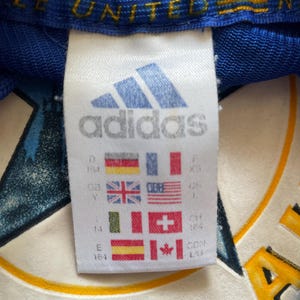 K&ouml;nnte beinhalten: Wei&szlig;es adidas-Etikett mit dem adidas-Logo und -Text. Das Etikett enth&auml;lt eine Gr&ouml;&szlig;entabelle mit verschiedenen L&auml;ndergr&ouml;&szlig;en. Das Etikett ist an einem blauen Shirt mit dem Text "LE UNITED" in Gelb befestigt.