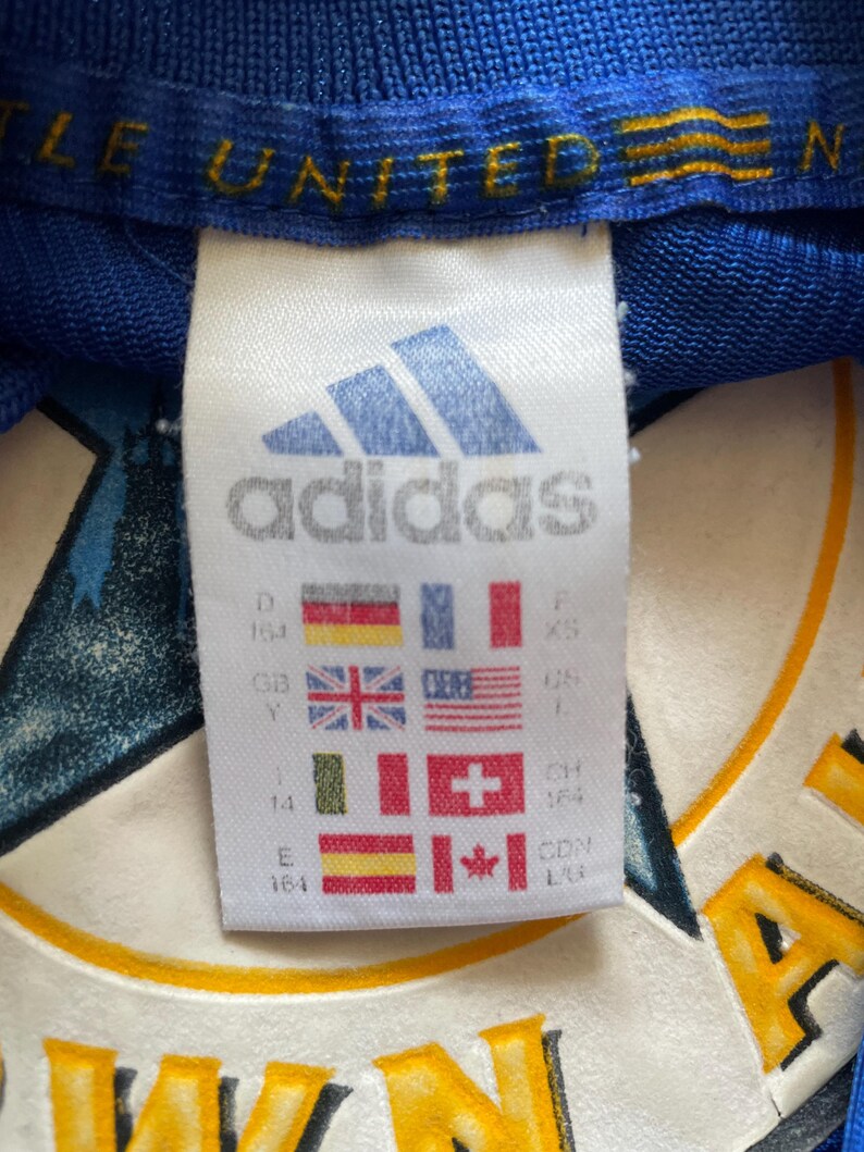 K&ouml;nnte beinhalten: Wei&szlig;es adidas-Etikett mit dem adidas-Logo und -Text. Das Etikett enth&auml;lt eine Gr&ouml;&szlig;entabelle mit verschiedenen L&auml;ndergr&ouml;&szlig;en. Das Etikett ist an einem blauen Shirt mit dem Text "LE UNITED" in Gelb befestigt.