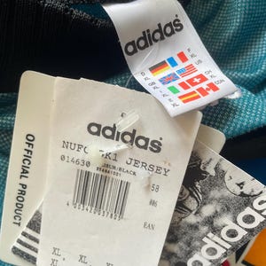 May include: Close-up of a teal and black Adidas jersey with attached tags. The tags display the Adidas logo, size information, and the text "NUFC K1 JERSEY". The jersey is an official product.