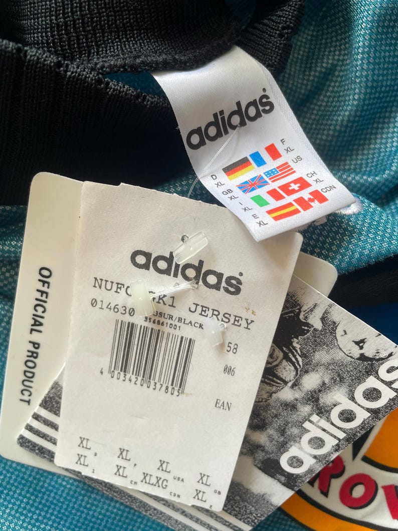 May include: Close-up of a teal and black Adidas jersey with attached tags. The tags display the Adidas logo, size information, and the text "NUFC K1 JERSEY". The jersey is an official product.