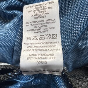 May include: A white label with text in English, German, and French, attached to blue fabric. The label details the brand, material composition, and care instructions. It also states the item was made in England. The fabric is a textured blue.