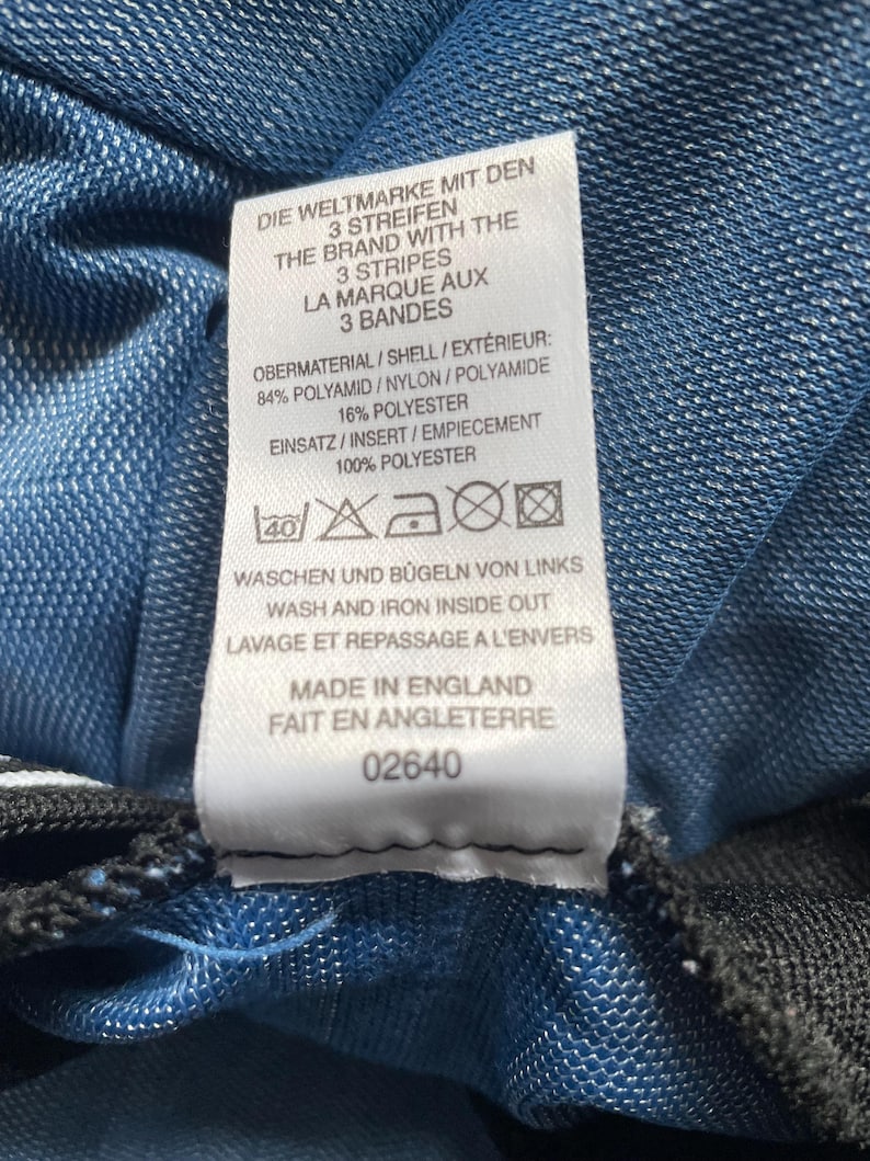 May include: A white label with text in English, German, and French, attached to blue fabric. The label details the brand, material composition, and care instructions. It also states the item was made in England. The fabric is a textured blue.