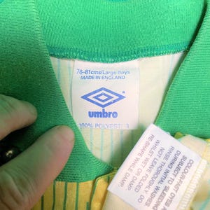 K&ouml;nnte beinhalten: Ein wei&szlig;es und gr&uuml;nes Fu&szlig;balltrikot mit gelben Streifen. Das Trikot hat ein wei&szlig;es Etikett mit dem Text "76-81cms/Large Boys MADE IN ENGLAND" und ein Logo f&uuml;r die Marke "umbro". Das Etikett sagt auch "100% POLYESTER". Es gibt ein zweites wei&szlig;es Etikett mit Pflegehinweisen.