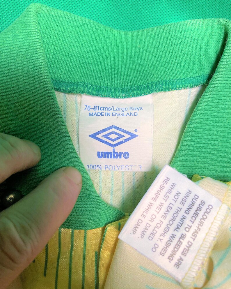 K&ouml;nnte beinhalten: Ein wei&szlig;es und gr&uuml;nes Fu&szlig;balltrikot mit gelben Streifen. Das Trikot hat ein wei&szlig;es Etikett mit dem Text "76-81cms/Large Boys MADE IN ENGLAND" und ein Logo f&uuml;r die Marke "umbro". Das Etikett sagt auch "100% POLYESTER". Es gibt ein zweites wei&szlig;es Etikett mit Pflegehinweisen.