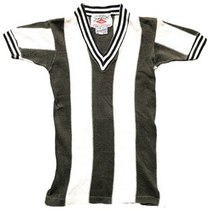 Puede incluir: Una camiseta vintage a rayas con cuello en V y mangas cortas. La camiseta es negra y blanca con una raya verde oscura en el centro. La camiseta tiene una peque&ntilde;a etiqueta con el texto "Champion" y "Made in USA" en la parte delantera.