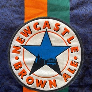 May include: Close-up of a navy blue fabric with orange and green stripes. A circular patch features a blue star and the words "NEWCASTLE BROWN ALE" in orange. The patch also includes a silhouette of a city skyline.