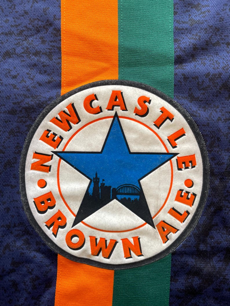 May include: Close-up of a navy blue fabric with orange and green stripes. A circular patch features a blue star and the words "NEWCASTLE BROWN ALE" in orange. The patch also includes a silhouette of a city skyline.