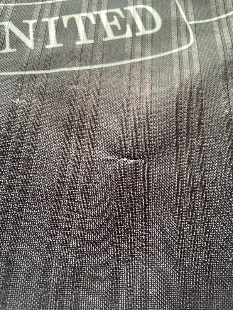 May include: Close-up of black fabric with a vertical ribbed texture. The word "UNITED" is visible in white block letters at the top. The fabric has a small tear.
