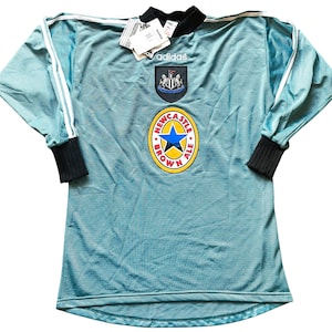K&ouml;nnte beinhalten: Hellblaues Adidas-Fu&szlig;balltrikot mit langen &Auml;rmeln. Es hat wei&szlig;e Streifen auf den &Auml;rmeln, einen schwarzen Kragen und B&uuml;ndchen sowie das Wappen und die Sponsorenlogos von Newcastle United. Ein Etikett ist am Kragen befestigt.
