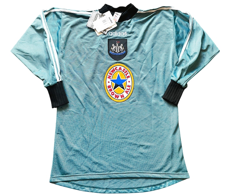 K&ouml;nnte beinhalten: Hellblaues Adidas-Fu&szlig;balltrikot mit langen &Auml;rmeln. Es hat wei&szlig;e Streifen auf den &Auml;rmeln, einen schwarzen Kragen und B&uuml;ndchen sowie das Wappen und die Sponsorenlogos von Newcastle United. Ein Etikett ist am Kragen befestigt.
