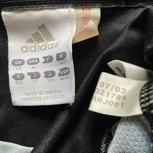 May include: A white tag with black text that reads "adidas" and size information in UK, USA, F, D, J, and CON sizes. The tag also says "Made in Tunisia" and "Fabrique en Tunisie". A white tag with black text that reads "07/03 021785 AHJ001".