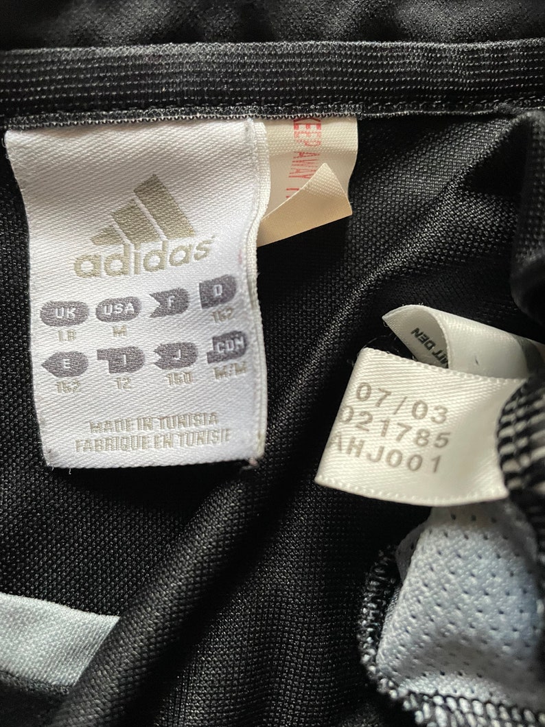 May include: A white tag with black text that reads "adidas" and size information in UK, USA, F, D, J, and CON sizes. The tag also says "Made in Tunisia" and "Fabrique en Tunisie". A white tag with black text that reads "07/03 021785 AHJ001".