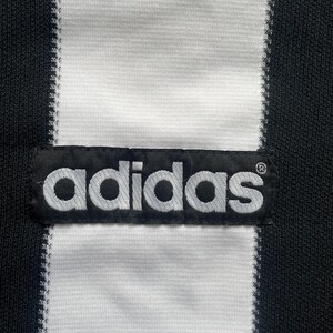 May include: Close-up of a black and white striped fabric with the Adidas logo patch. The Adidas logo is in white, and the fabric has a textured appearance.