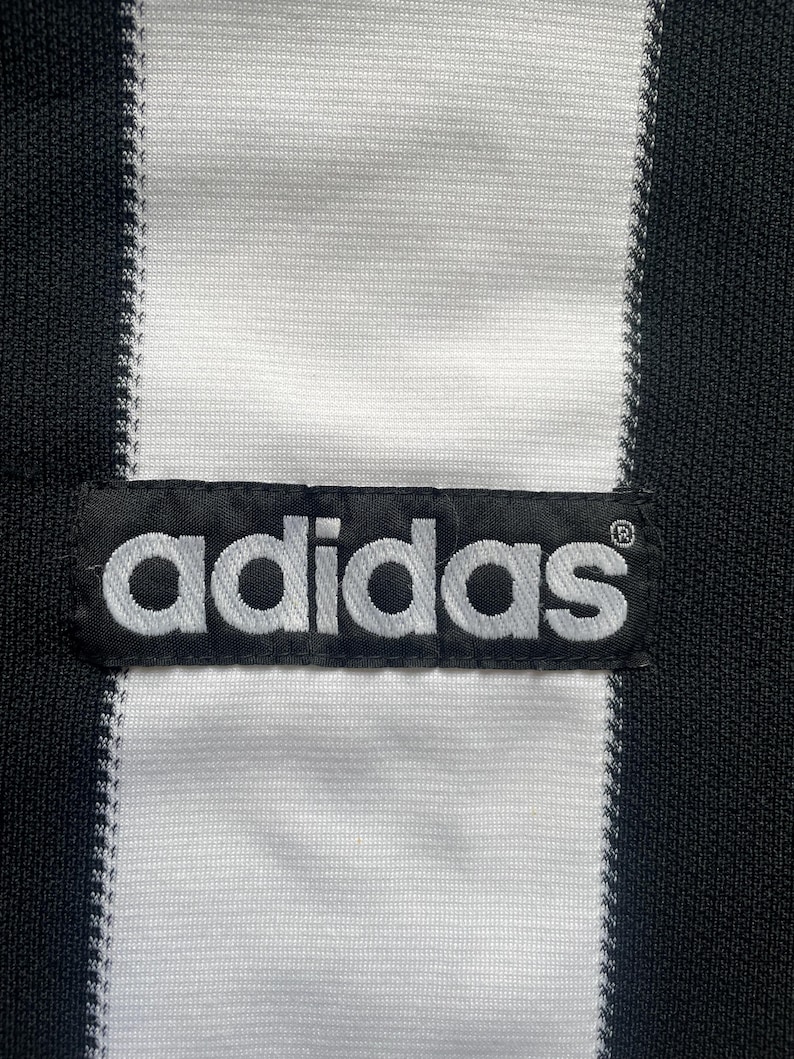 May include: Close-up of a black and white striped fabric with the Adidas logo patch. The Adidas logo is in white, and the fabric has a textured appearance.