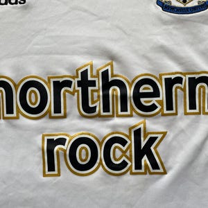 Pode incluir: Camisola de futebol branca com as palavras "northern rock" em preto com contornos dourados. O log&oacute;tipo da Adidas &eacute; vis&iacute;vel no canto superior esquerdo. O bras&atilde;o do Newcastle United est&aacute; no canto superior direito.