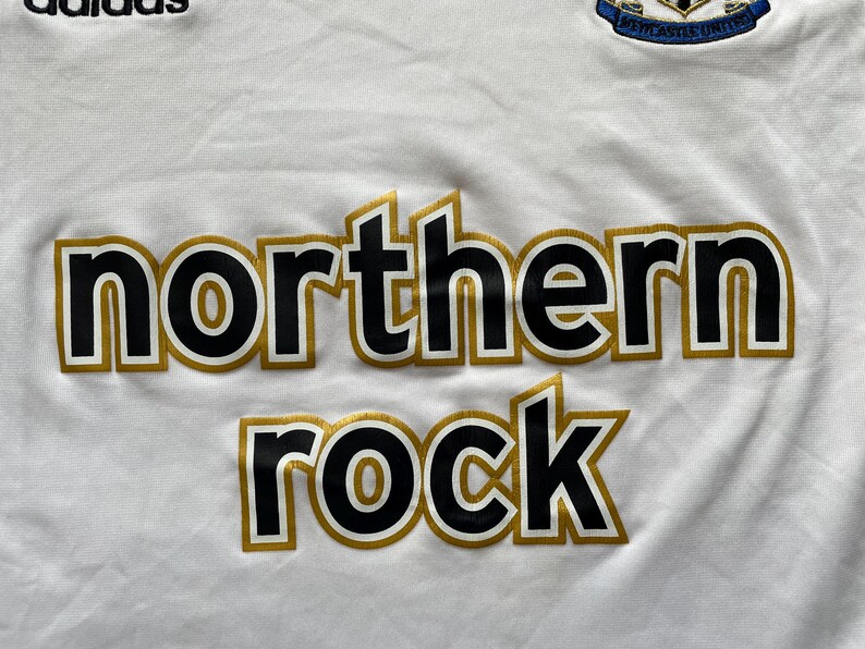 Pode incluir: Camisola de futebol branca com as palavras "northern rock" em preto com contornos dourados. O log&oacute;tipo da Adidas &eacute; vis&iacute;vel no canto superior esquerdo. O bras&atilde;o do Newcastle United est&aacute; no canto superior direito.