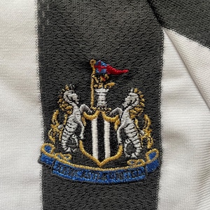 May include: Detail of a black and white striped fabric with an embroidered crest. The crest displays a shield with a black and white vertical pattern, flanked by two seahorses, and a banner with text.