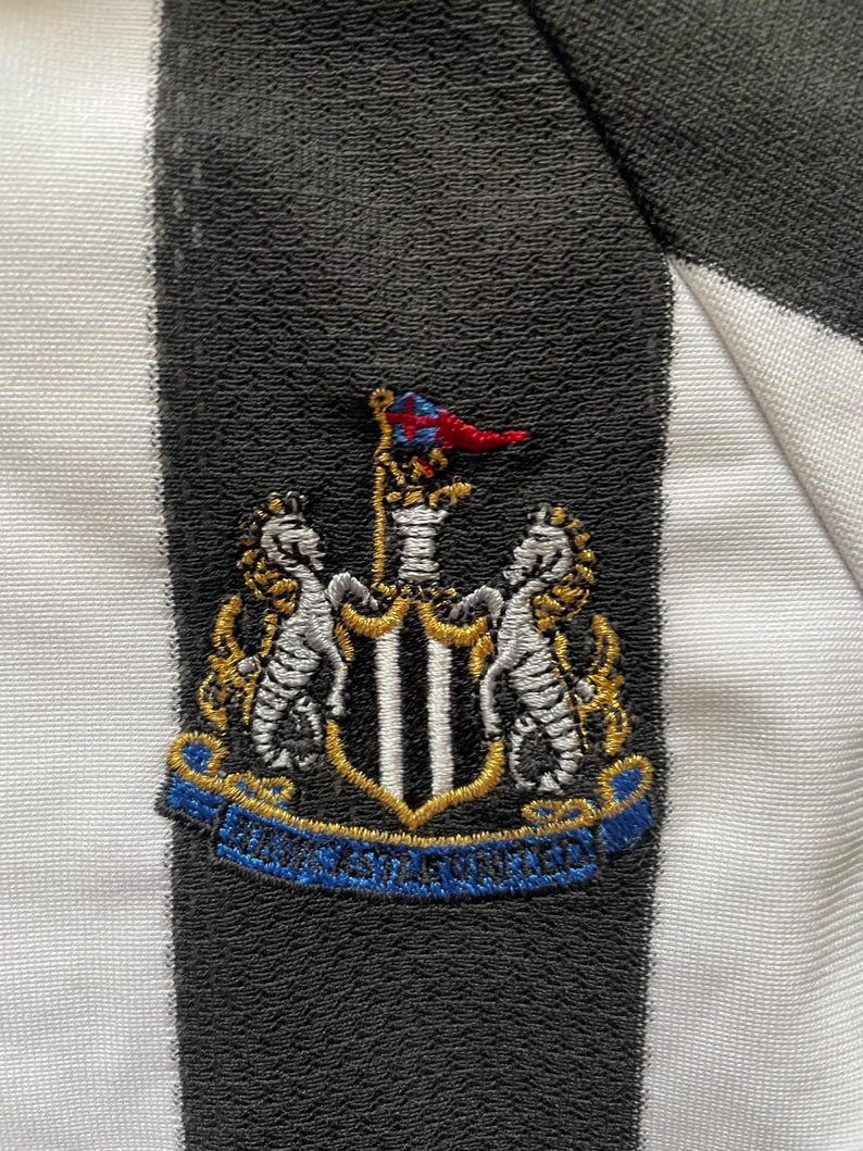 May include: Detail of a black and white striped fabric with an embroidered crest. The crest displays a shield with a black and white vertical pattern, flanked by two seahorses, and a banner with text.