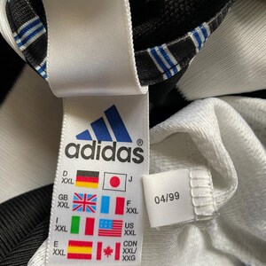 May include: White and black adidas shirt with a size tag that shows the size in different countries. The tag also includes the date 04/99.