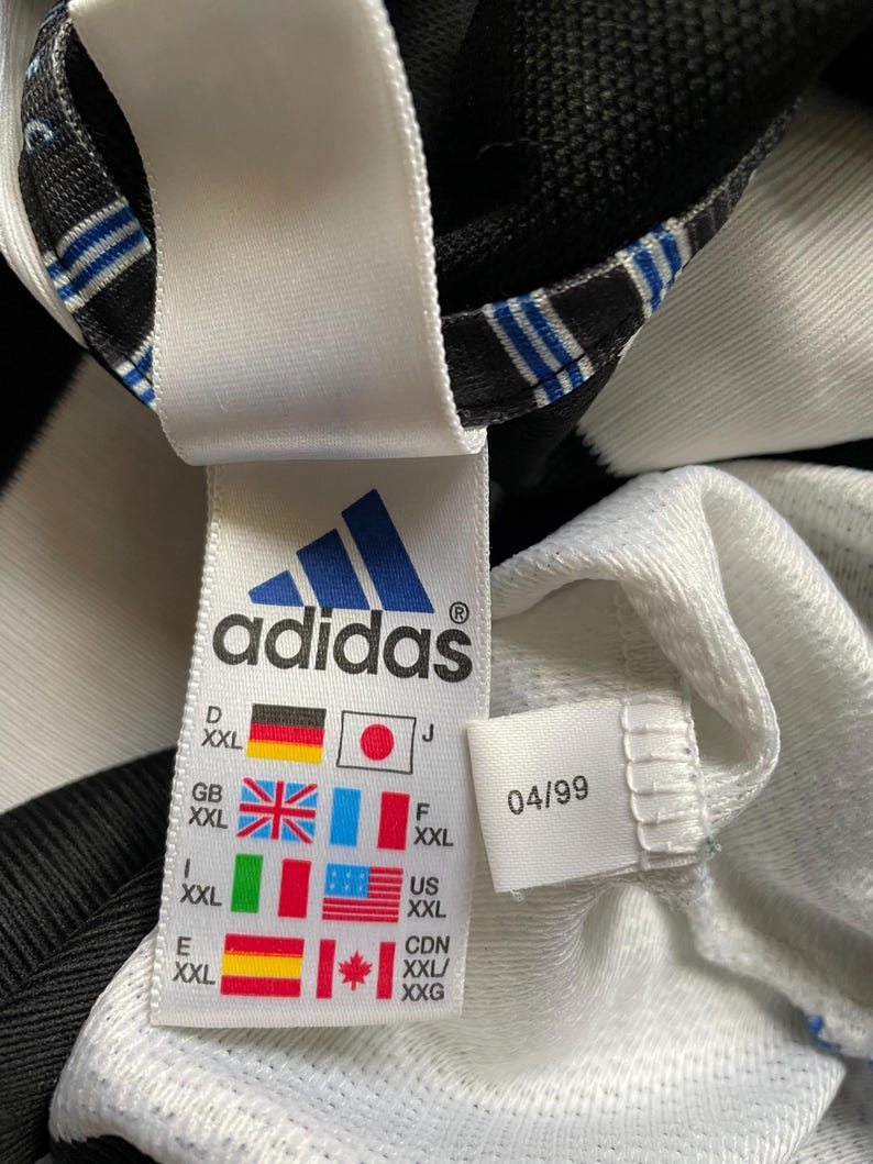 May include: White and black adidas shirt with a size tag that shows the size in different countries. The tag also includes the date 04/99.