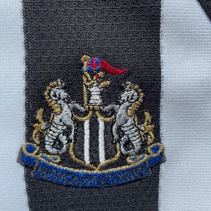 Pode incluir: Um remendo bordado em preto e branco com um bras&atilde;o que apresenta dois cavalos-marinhos prateados flanqueando um escudo com duas faixas verticais pretas. A crista &eacute; coroada com uma bandeira vermelha e azul e uma fita dourada com o texto "Newcastle United" em azul.