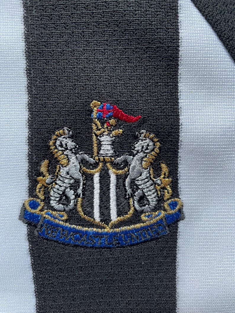 Pode incluir: Um remendo bordado em preto e branco com um bras&atilde;o que apresenta dois cavalos-marinhos prateados flanqueando um escudo com duas faixas verticais pretas. A crista &eacute; coroada com uma bandeira vermelha e azul e uma fita dourada com o texto "Newcastle United" em azul.
