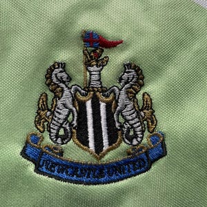 May include: A green and white soccer jersey with the Newcastle United crest embroidered on the front. The crest features a black and white shield with two gold seahorses on either side. The crest also includes a blue ribbon with the words "Newcastle United" written in white.