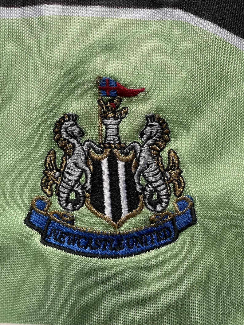May include: A green and white soccer jersey with the Newcastle United crest embroidered on the front. The crest features a black and white shield with two gold seahorses on either side. The crest also includes a blue ribbon with the words "Newcastle United" written in white.