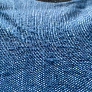 May include: Close-up of blue denim fabric with a textured surface. The fabric has a faded appearance, with a darker shade at the bottom and a lighter shade at the top. A circular patch with the word "TOWN" is visible.