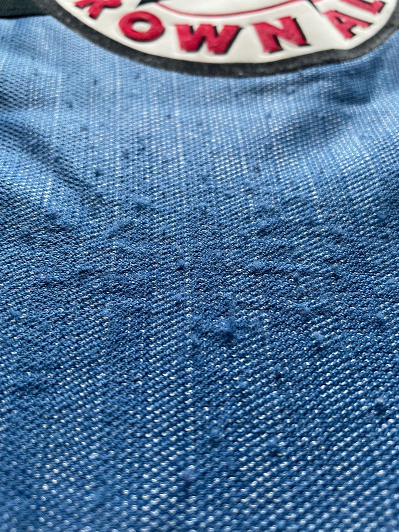 May include: Close-up of blue denim fabric with a textured surface. The fabric has a faded appearance, with a darker shade at the bottom and a lighter shade at the top. A circular patch with the word "TOWN" is visible.