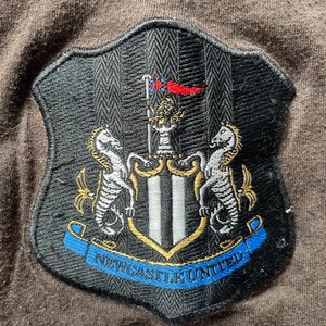 Puede incluir: Parche bordado con el escudo del Newcastle United. El escudo, con forma de escudo, tiene una parte superior a rayas blancas y negras y una pancarta azul con el texto "NEWCASTLE UNITED". El escudo incluye dos caballitos de mar y un emblema central.
