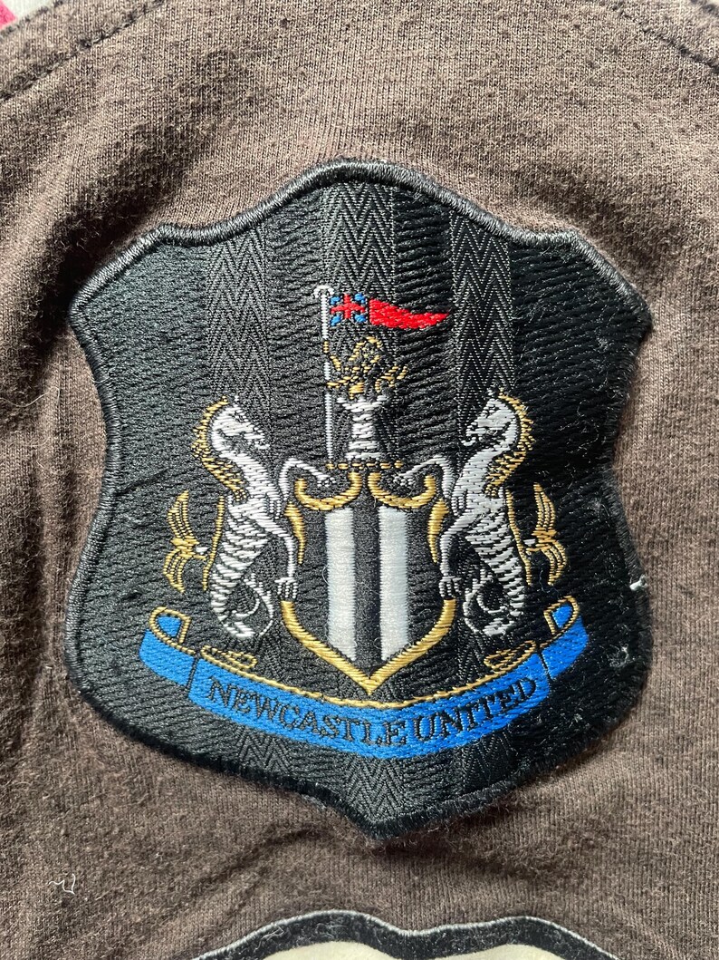 Puede incluir: Parche bordado con el escudo del Newcastle United. El escudo, con forma de escudo, tiene una parte superior a rayas blancas y negras y una pancarta azul con el texto "NEWCASTLE UNITED". El escudo incluye dos caballitos de mar y un emblema central.