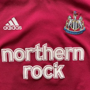 Pu&ograve; includere: Una maglia da calcio rossa con il logo Adidas, lo stemma del Newcastle United e le parole "northern rock" in bianco.