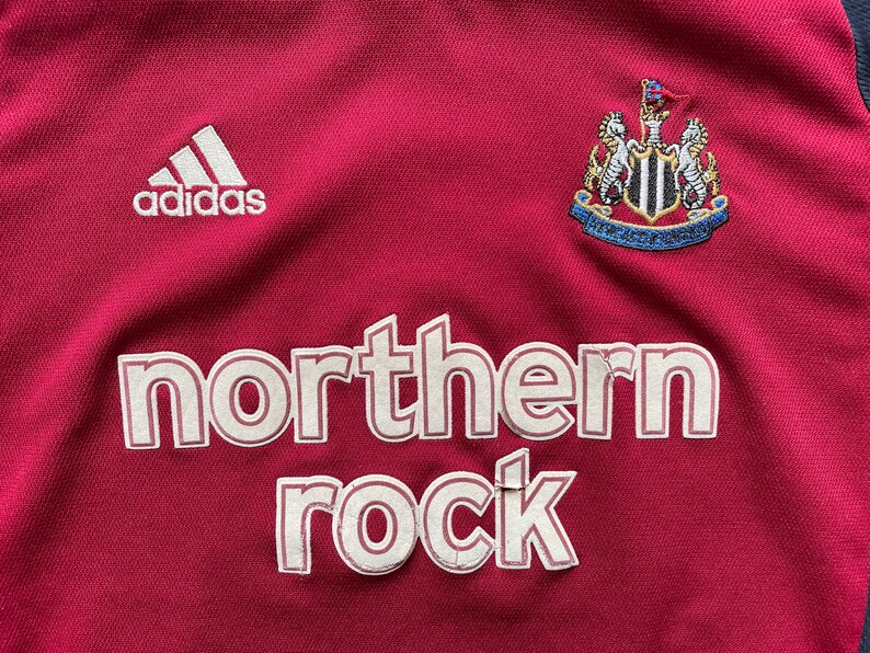 Pu&ograve; includere: Una maglia da calcio rossa con il logo Adidas, lo stemma del Newcastle United e le parole "northern rock" in bianco.