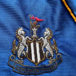 K&ouml;nnte beinhalten: Ein blaues Fu&szlig;balltrikot mit dem gestickten Wappen von Newcastle United auf der Vorderseite. Das Wappen zeigt einen schwarz-wei&szlig;en Schild mit zwei goldenen Seepferdchen auf jeder Seite. Das Wappen enth&auml;lt auch ein goldenes Banner mit den Worten "Newcastle United" in Schwarz.