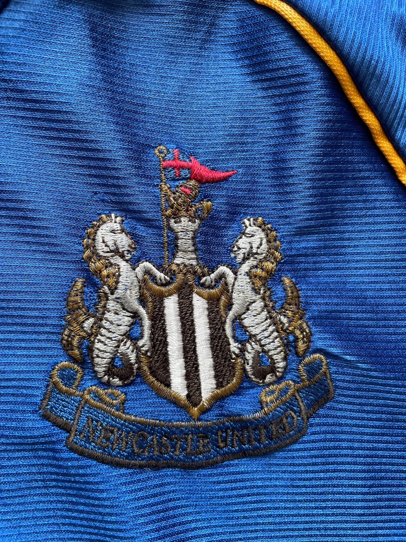 K&ouml;nnte beinhalten: Ein blaues Fu&szlig;balltrikot mit dem gestickten Wappen von Newcastle United auf der Vorderseite. Das Wappen zeigt einen schwarz-wei&szlig;en Schild mit zwei goldenen Seepferdchen auf jeder Seite. Das Wappen enth&auml;lt auch ein goldenes Banner mit den Worten "Newcastle United" in Schwarz.