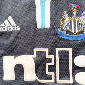 Op de afbeelding: Zwart voetbaltenue met een wit Adidas-logo, een wit "nt!"-sponsorlogo en een Newcastle United-wapen.