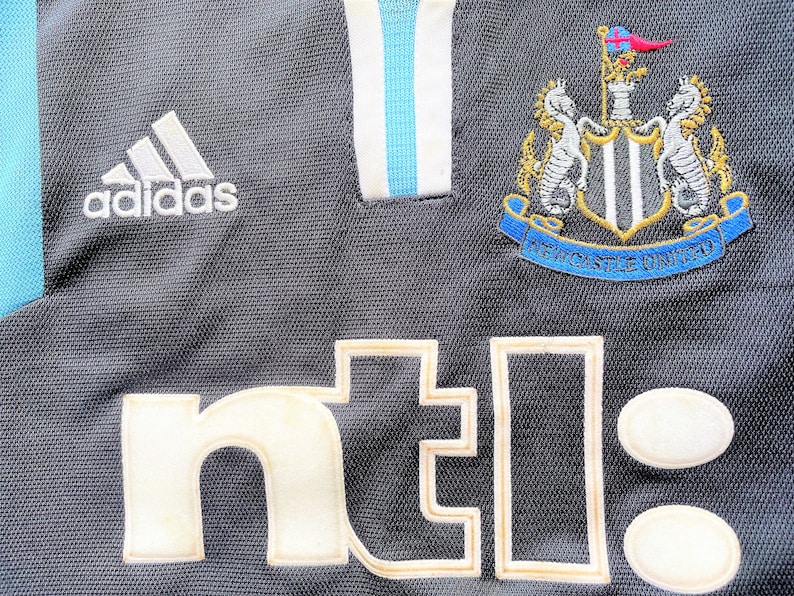 Op de afbeelding: Zwart voetbaltenue met een wit Adidas-logo, een wit "nt!"-sponsorlogo en een Newcastle United-wapen.