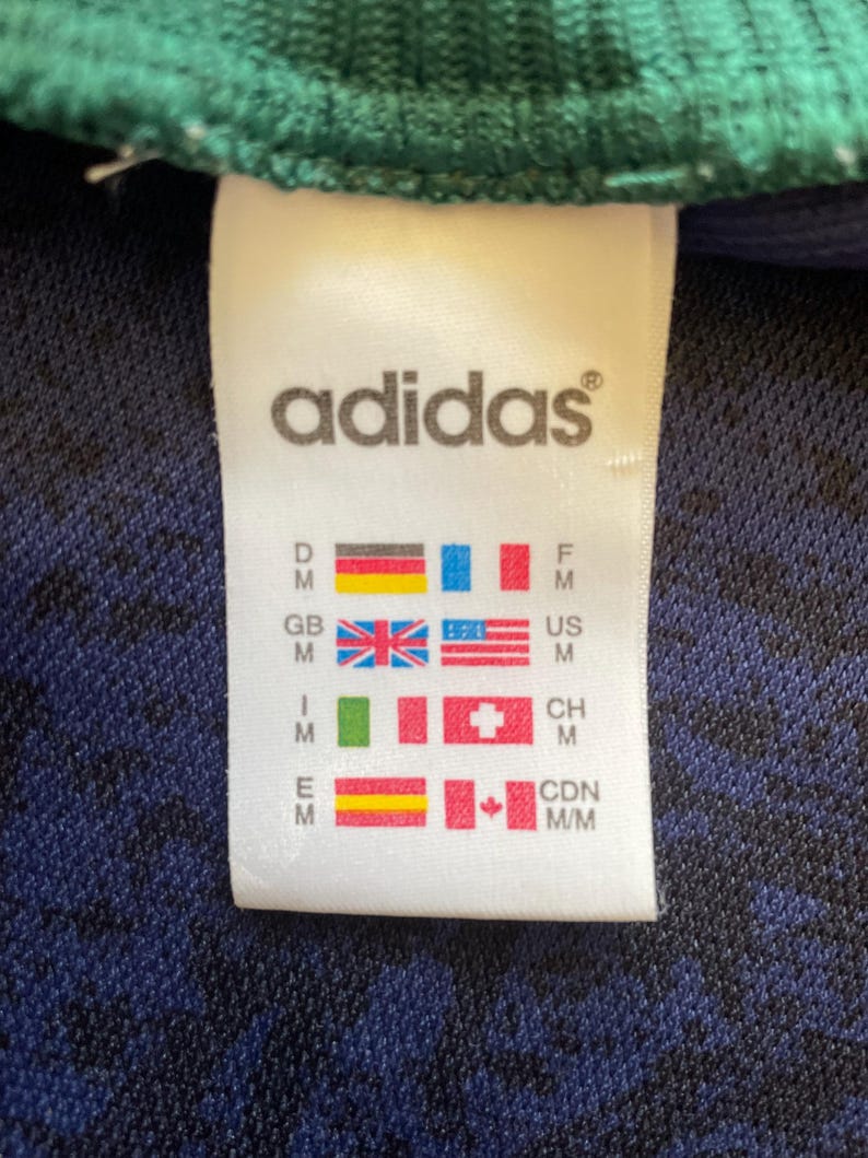 May include: Close-up of a white Adidas tag with the brand name and various country flags. The tag is on a dark blue and black textured fabric, with a green ribbed edge visible at the top.