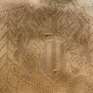 May include: Close-up of a textured fabric in a warm, golden-brown hue. The material features a repeating pattern of small, raised squares and subtle geometric designs, creating a tactile and visually interesting surface. The fabric appears to be woven.