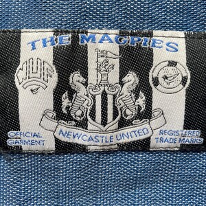 May include: A rectangular patch with black and white vertical stripes, featuring the text "THE MAGPIES" in blue. The patch includes the Newcastle United crest, text "OFFICIAL GARMENT" and "REGISTERED TRADE MARKS".