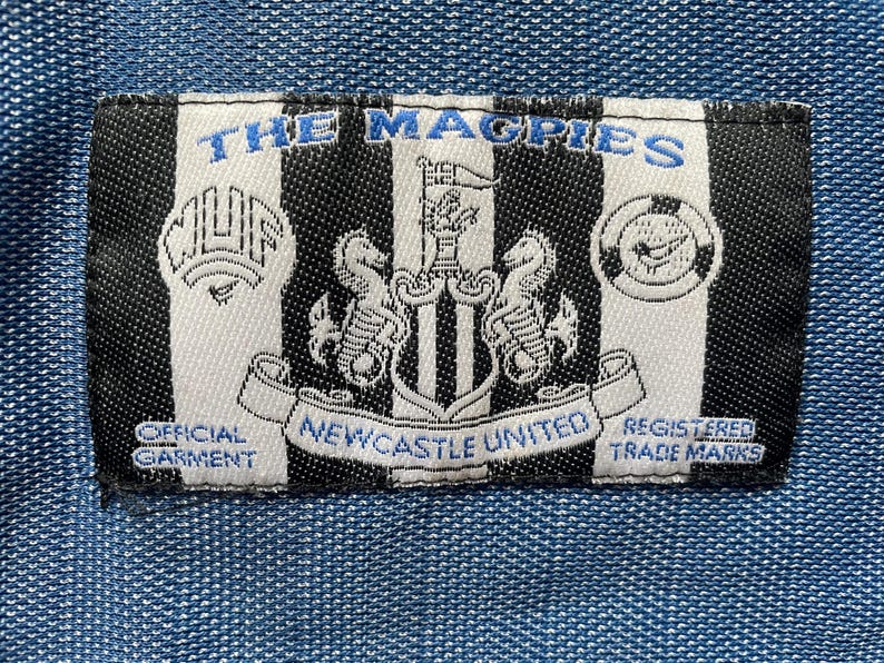 May include: A rectangular patch with black and white vertical stripes, featuring the text "THE MAGPIES" in blue. The patch includes the Newcastle United crest, text "OFFICIAL GARMENT" and "REGISTERED TRADE MARKS".