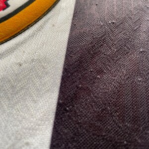 May include: Close-up of a fabric with a white, gray, and dark brown color scheme. The fabric has a textured appearance with small, raised bumps. A portion of a circular patch with red text and a yellow border is visible.
