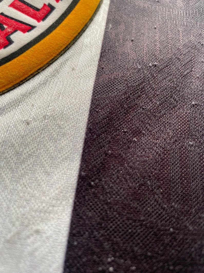 May include: Close-up of a fabric with a white, gray, and dark brown color scheme. The fabric has a textured appearance with small, raised bumps. A portion of a circular patch with red text and a yellow border is visible.