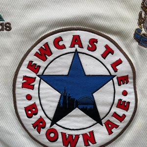 May include: Detail of a white fabric with a circular embroidered patch. The patch reads "NEWCASTLE BROWN ALE" in red, encircling a blue star with a city skyline. An Adidas logo is also visible.