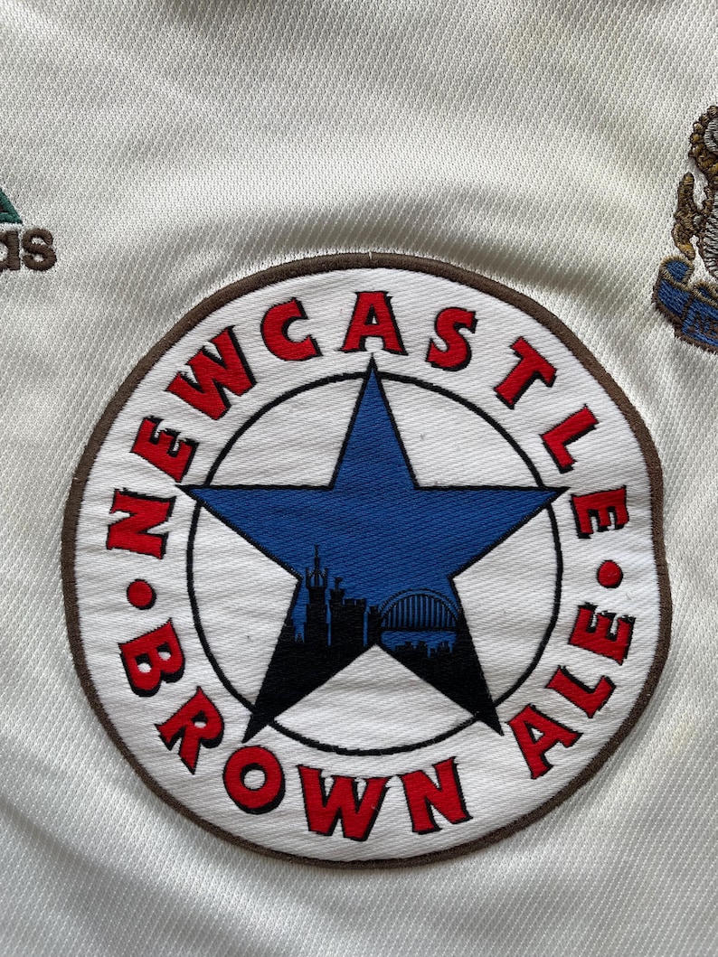 May include: Detail of a white fabric with a circular embroidered patch. The patch reads "NEWCASTLE BROWN ALE" in red, encircling a blue star with a city skyline. An Adidas logo is also visible.