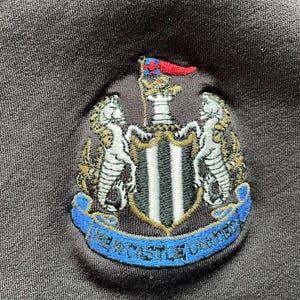 May include: Black embroidered patch with a crest featuring a shield with black and white stripes, two gold lions, and a gold crown. The crest is surrounded by a blue ribbon with the text "Newcastle United" in white.