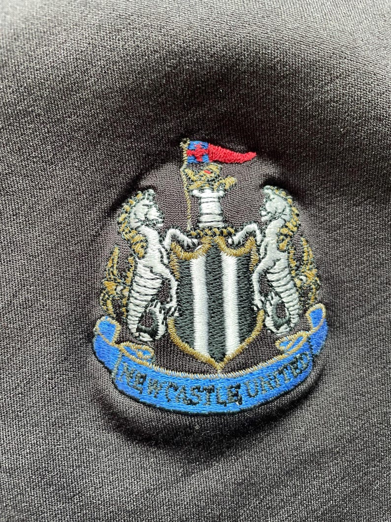May include: Black embroidered patch with a crest featuring a shield with black and white stripes, two gold lions, and a gold crown. The crest is surrounded by a blue ribbon with the text "Newcastle United" in white.